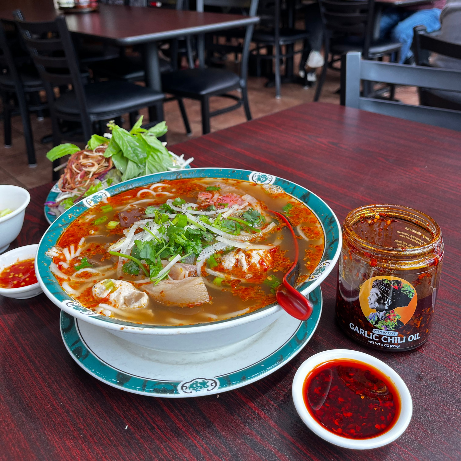 A bowl of hot bun bo hue noodle soup with green herbs on top, served on a dark wooden restaurant table with a jar of Mien Market garlic chili oil and a small sauce plate of garlic chili oil for dipping. The background shows a busy restaurant interior with tables and seated people.