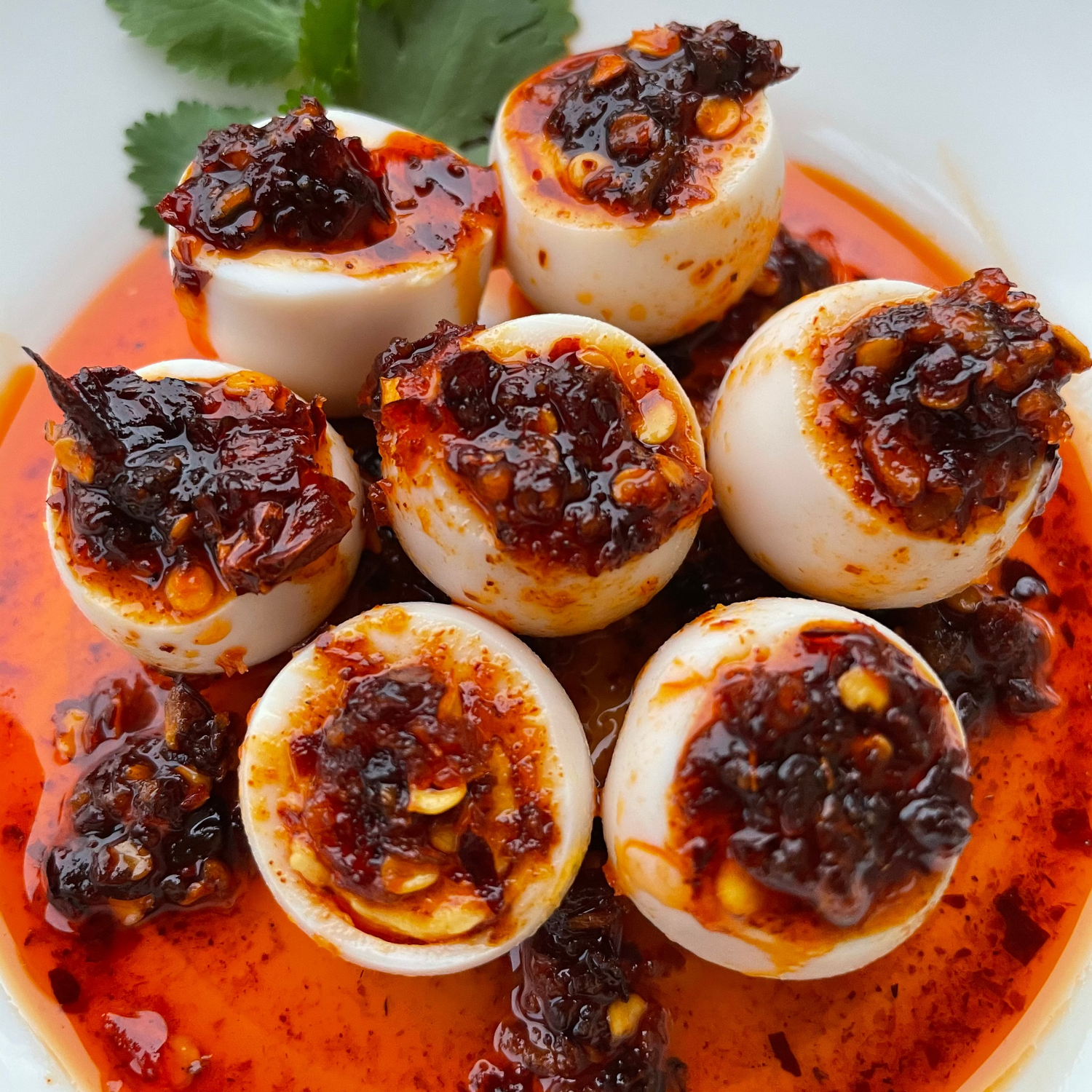 Six halved quail eggs filled with Mien Market Garlic chili Oil garnished with cilantro on a white plate.