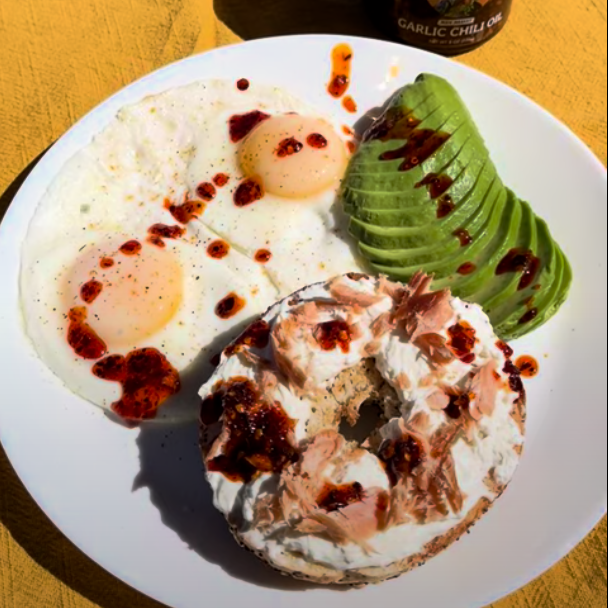 A breakfast plate with two fried eggs, a bagel topped with smoked salmon, cream cheese and Mien Market Garlic Chili Oil along with a small jar of garlic chili oil on a yellow cloth outdoors with grass in the background.